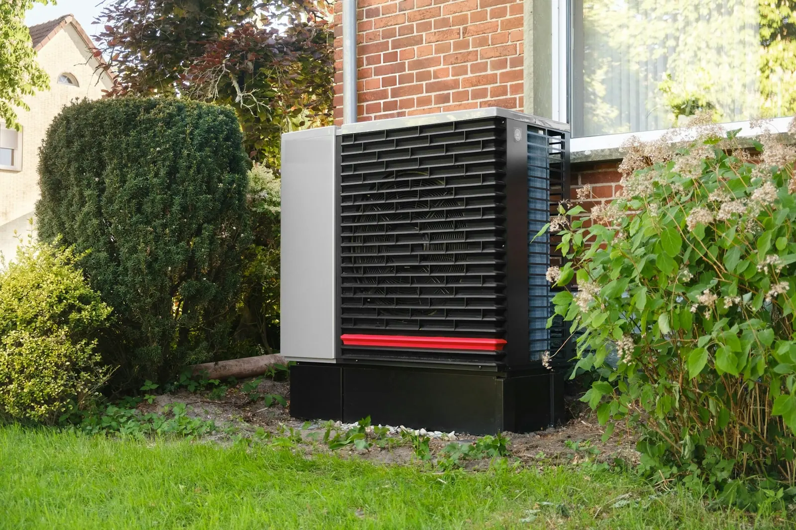 Een warmtepomp buiten naast een bakstenen huis met een tuin